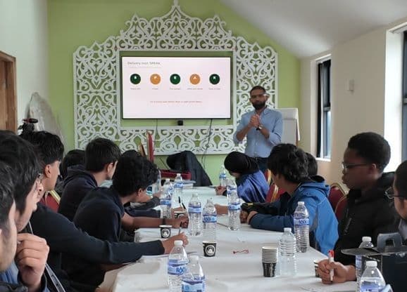 Imran mid-sentence in front of a screen, running a public speaking session for a room of teenagers seated around a long table.
